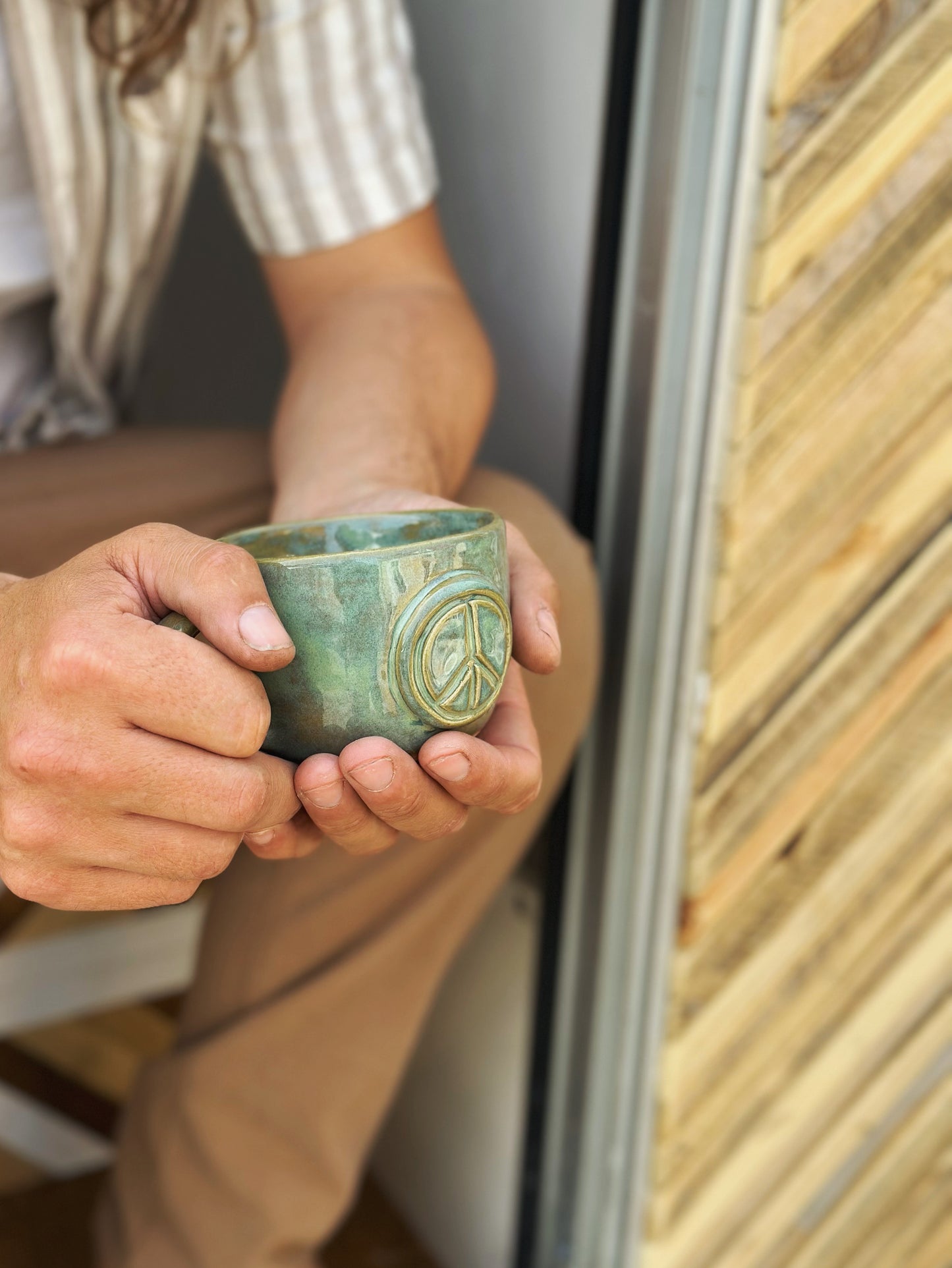 Man sitting in the opening of a camper van, holding a green ceramic mug in his hands decorated with a peace sign.