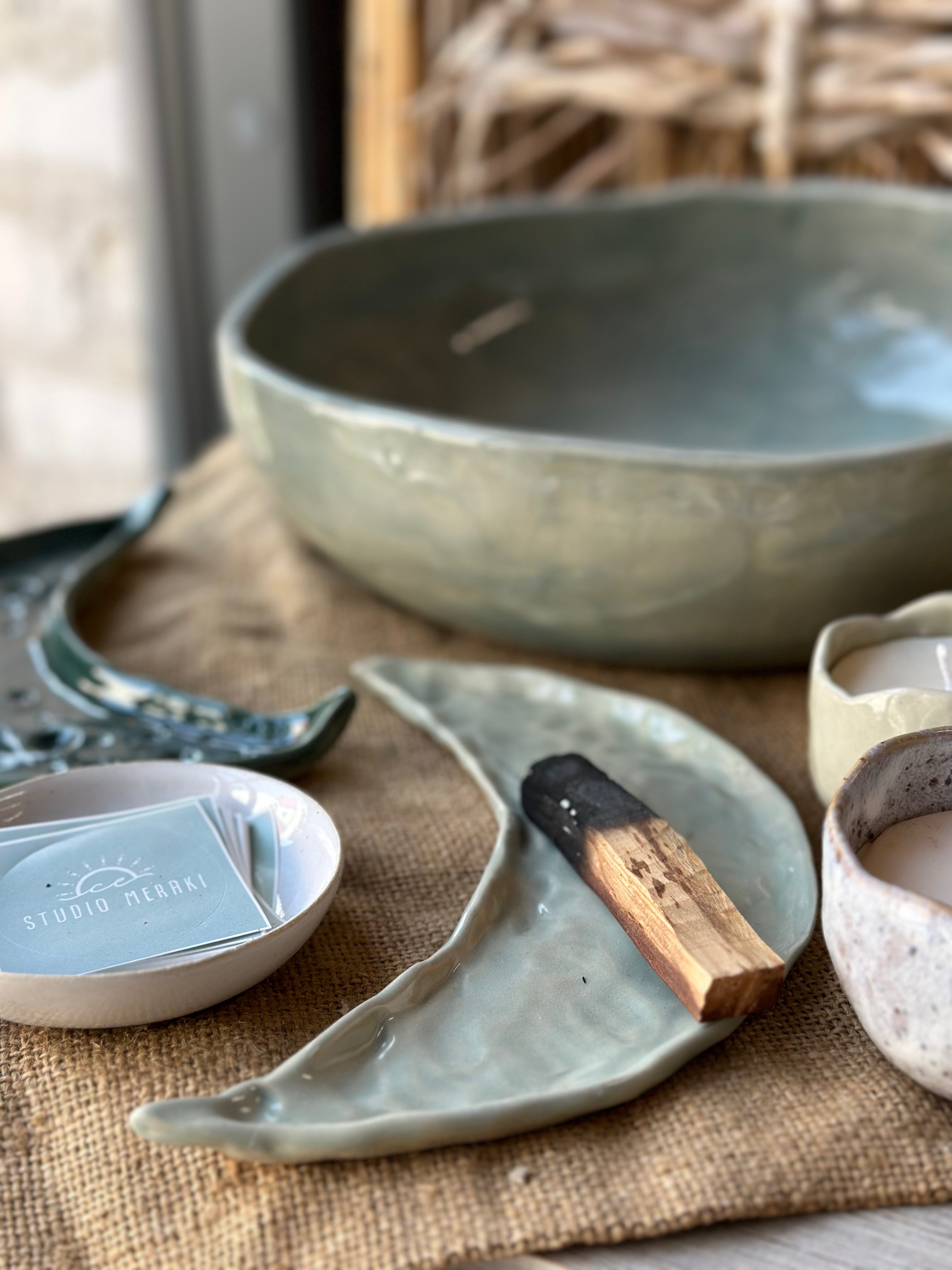 Close up from a pottery shop showing a large bowl in the background and a moon shaped ceramic tray with Palo Santo insence in the foreground.