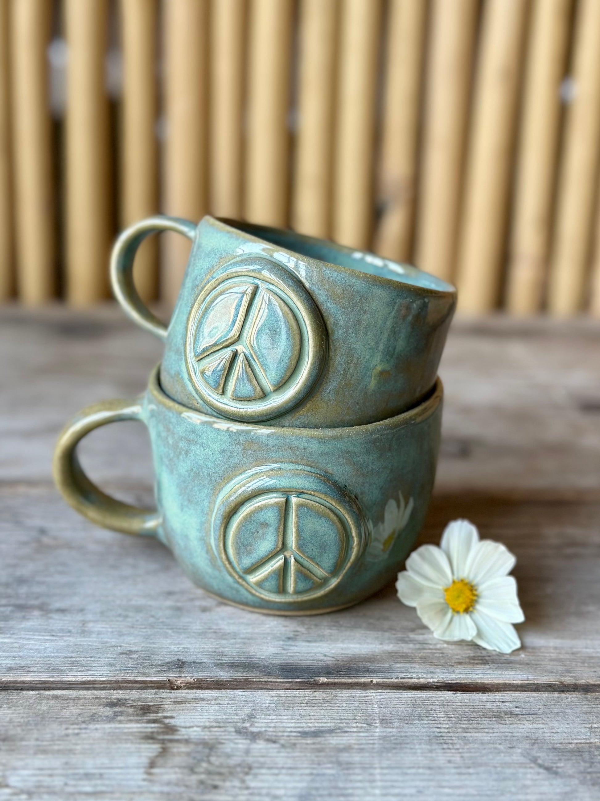 Two green ceramic cup with handle, decorated with a peace sign. Stacked on top of each other. Flower on the side.