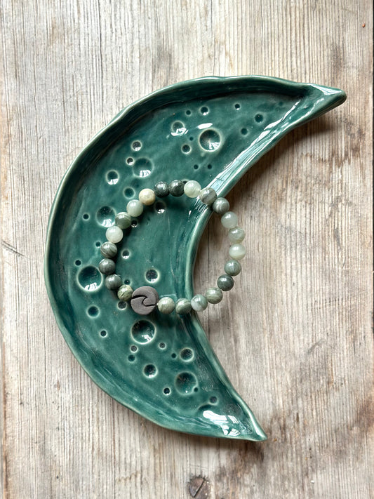 Moon shaped tray in dark green colour, on the tray lays a chrystal bracelet decorated with a ceramic pearl in the shape of a wave.