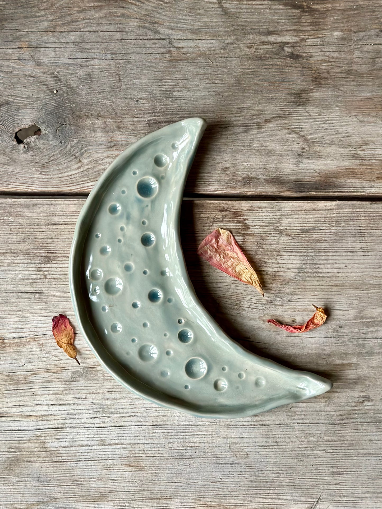 Moon shaped decorative tray in grey blue color. Around the tray lays dried flowers.