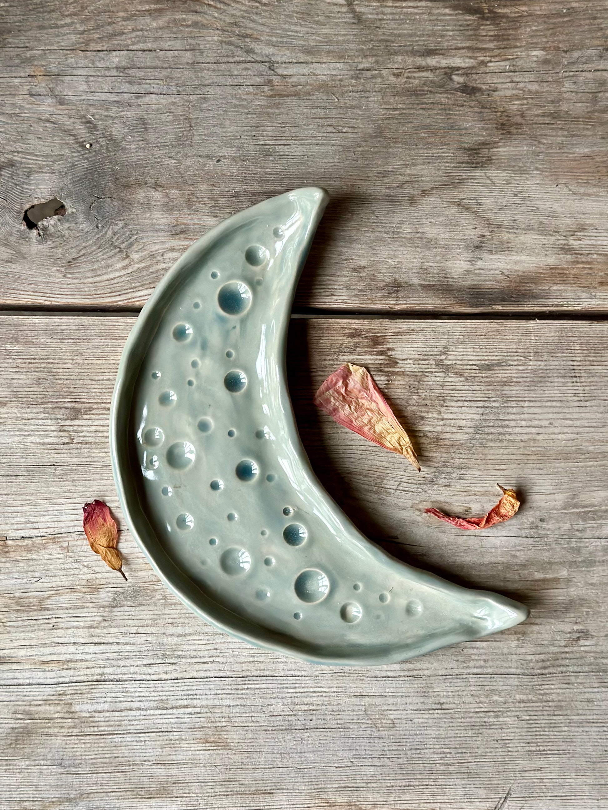 Moon shaped decorative tray in grey blue color. Around the tray lays dried flowers.