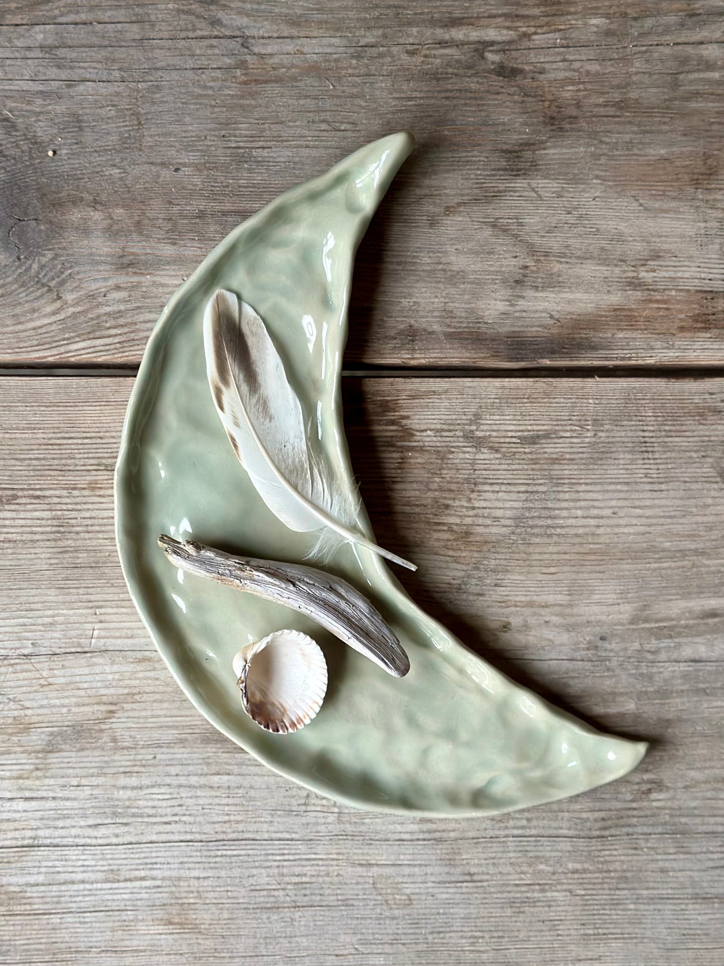 Moon shaped decorative tray in light aqua green color. On the tray lays a shell, a drift wood stick and a feather.