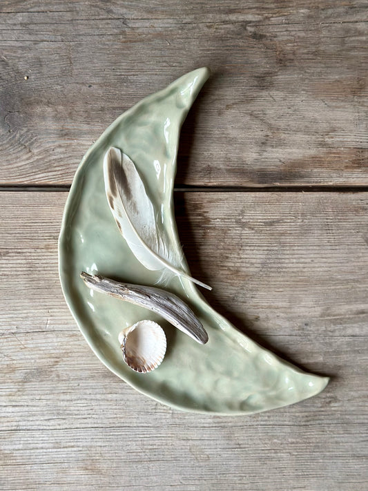 Moon shaped decorative tray in light aqua green color. On the tray lays a shell, a drift wood stick and a feather.