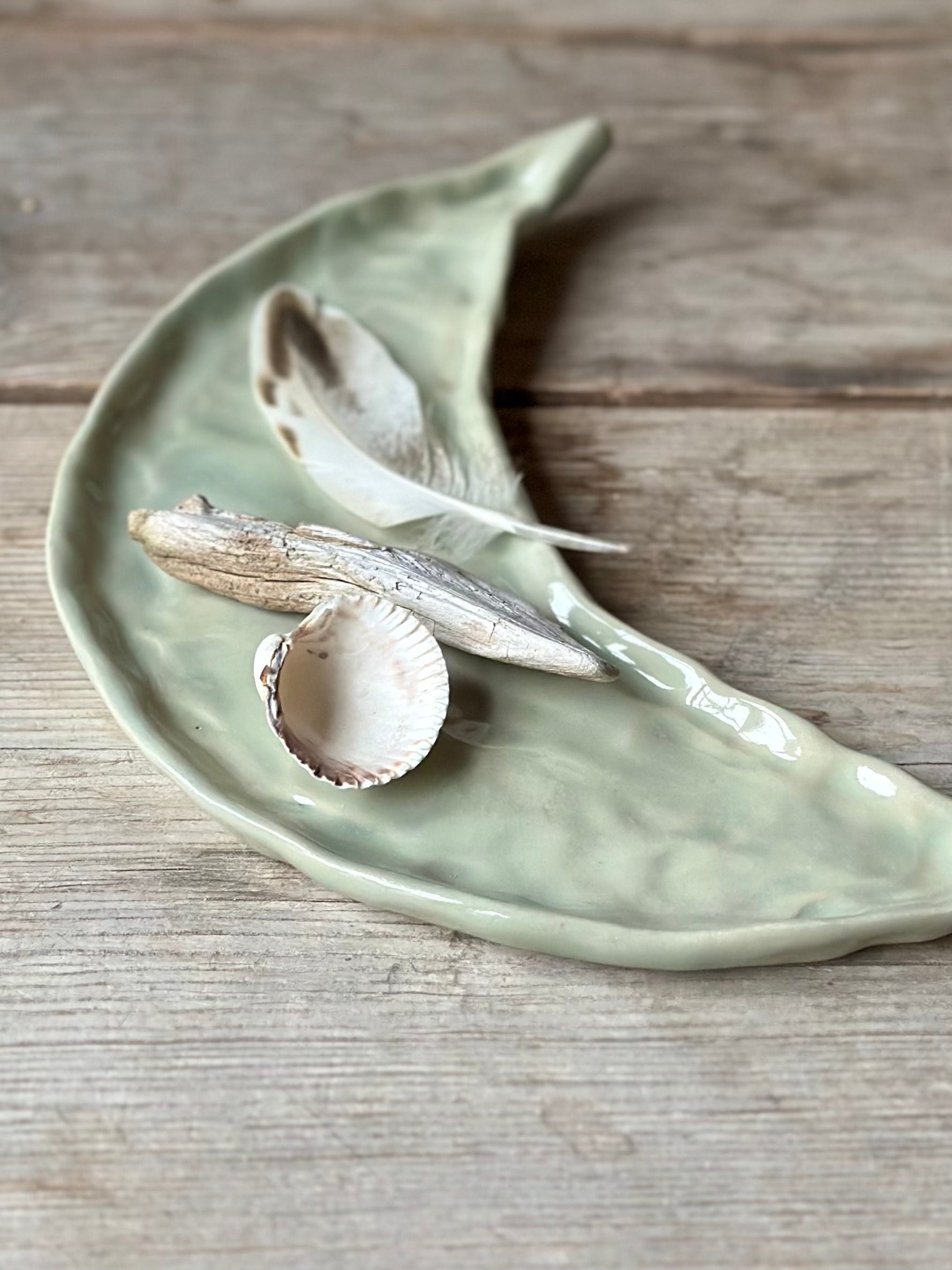 Moon shaped decorative tray in light aqua green color. On the tray lays a shell, a drift wood stick and a feather.