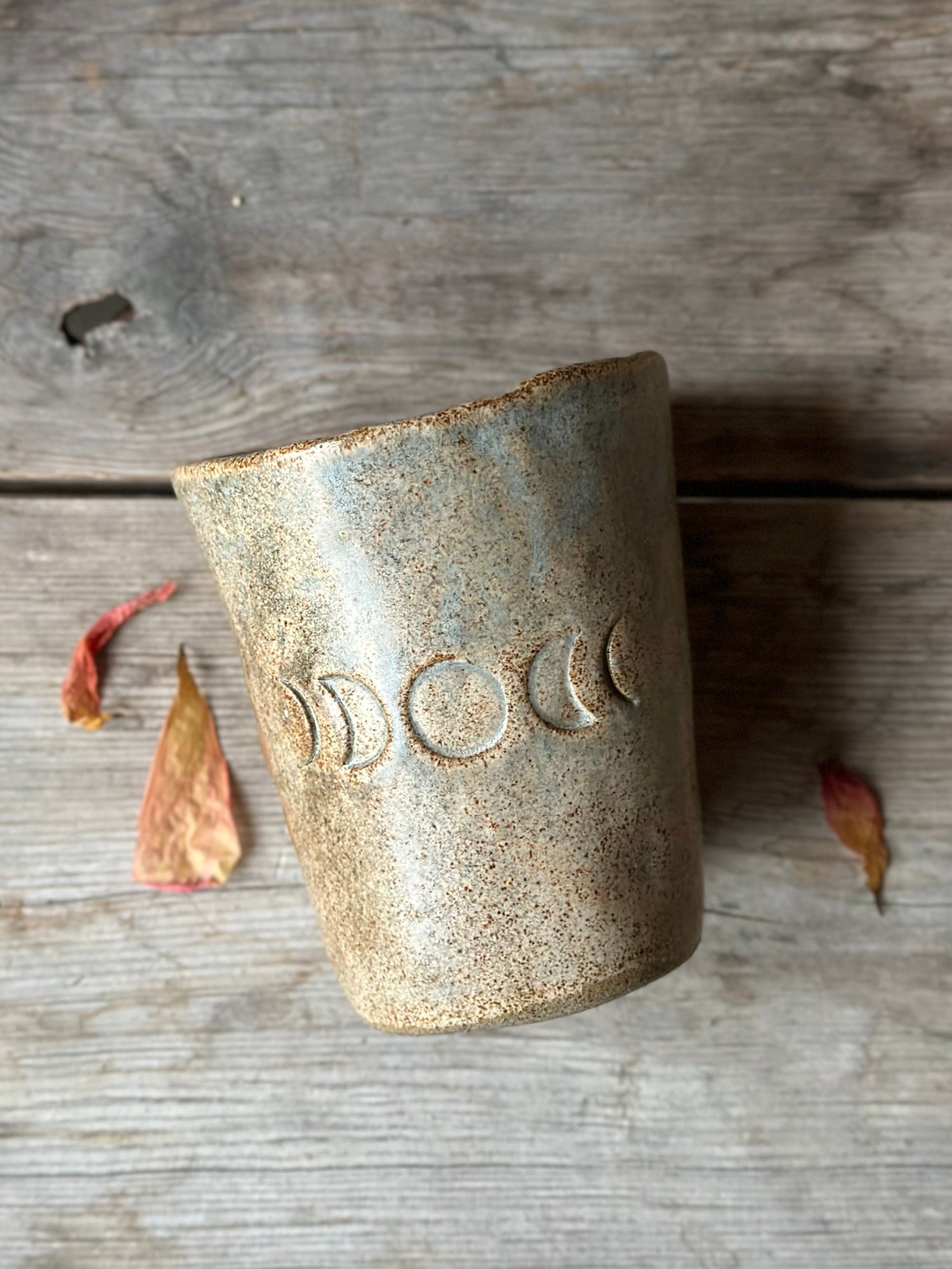 Beige grey ceramic cup laying on a grey old table. The cup is decorated with moon phases. Around the cup is dried flowers.