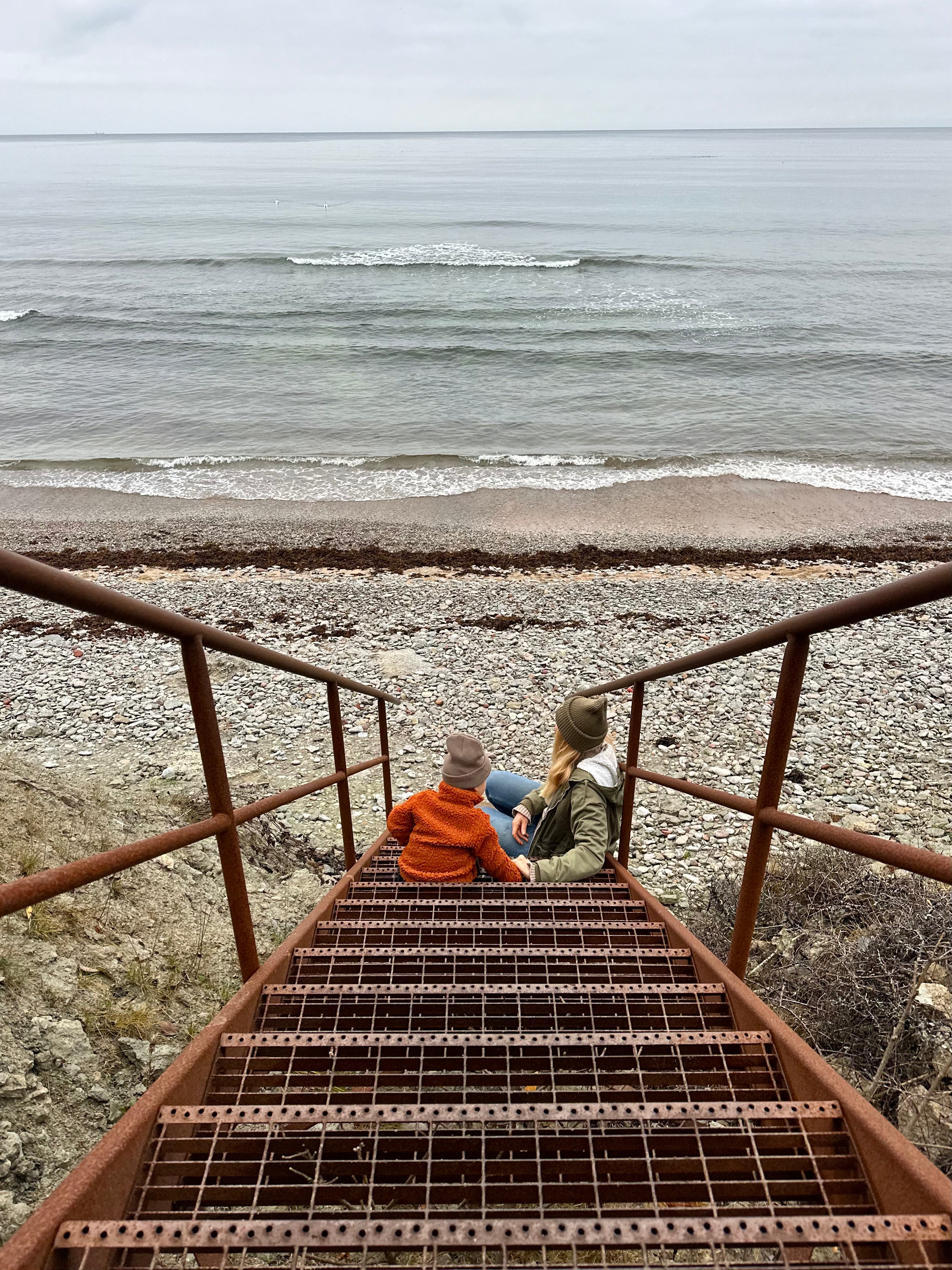 Woman and child dressed in winter clothes sitting on a rusty stairs down to a stone bech, looking out over the sea.