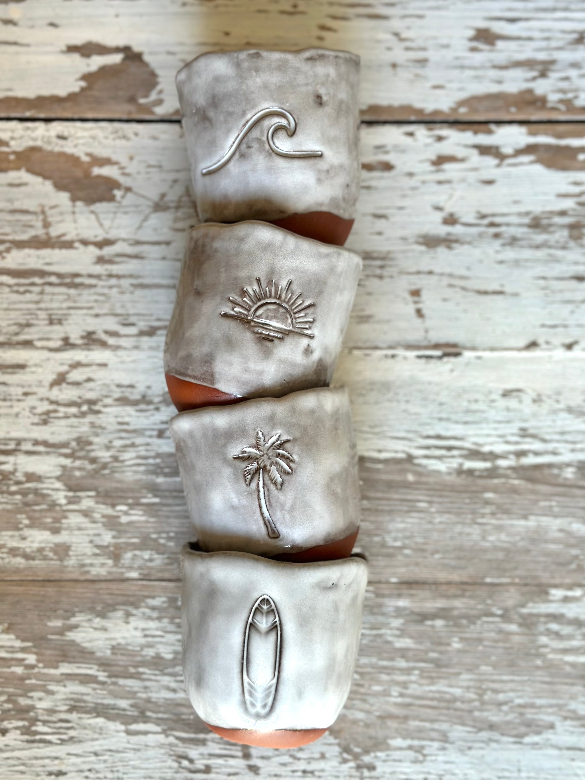 Stack of four ceramic cups laying on the table. They are made in brown clay and glazed in a light grey glaze. Decorated with different beach like symbols.