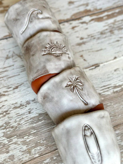 Four ceramic mugs stacked on a table. The mugs are decorated with beach vibe symbols like a wave, sunset, palm tree and surfboard.