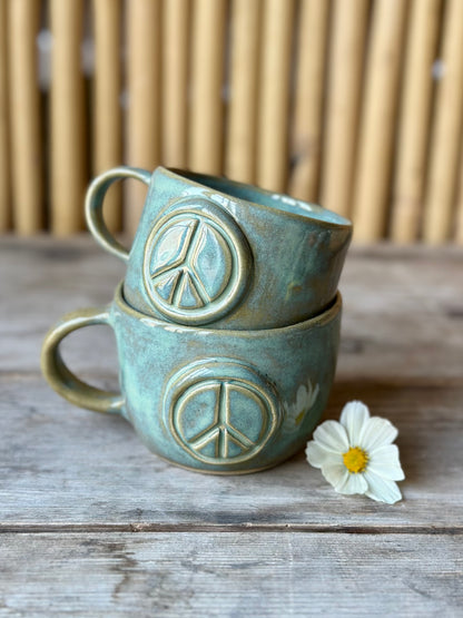 Two green ceramic cup with handle, decorated with a peace sign. Stacked on top of each other. Flower on the side.