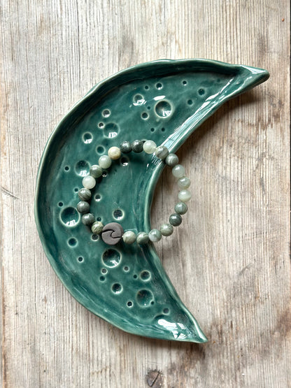 Moon shaped tray in dark green colour, on the tray lays a chrystal bracelet decorated with a ceramic pearl in the shape of a wave.