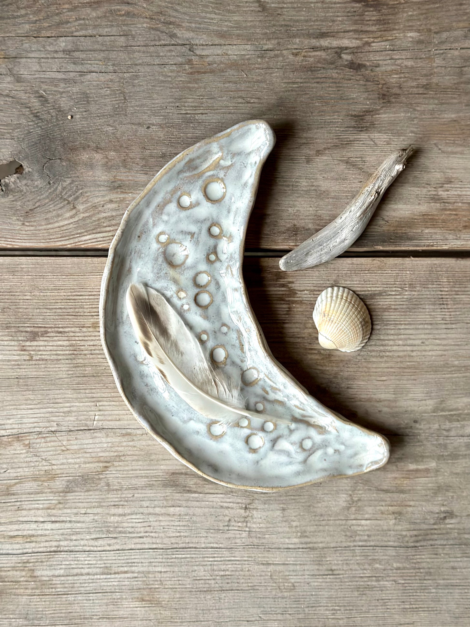 Moon shaped decorative tray in light beige and white color. On the tray lays a feather and around it is a shell and some drift wood.