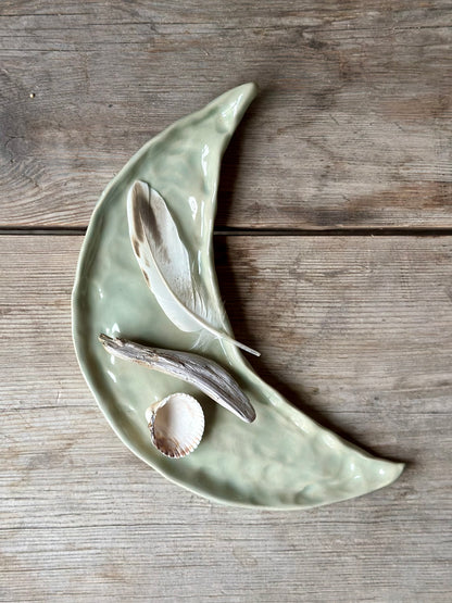 Moon shaped decorative tray in light aqua green color. On the tray lays a shell, a drift wood stick and a feather.