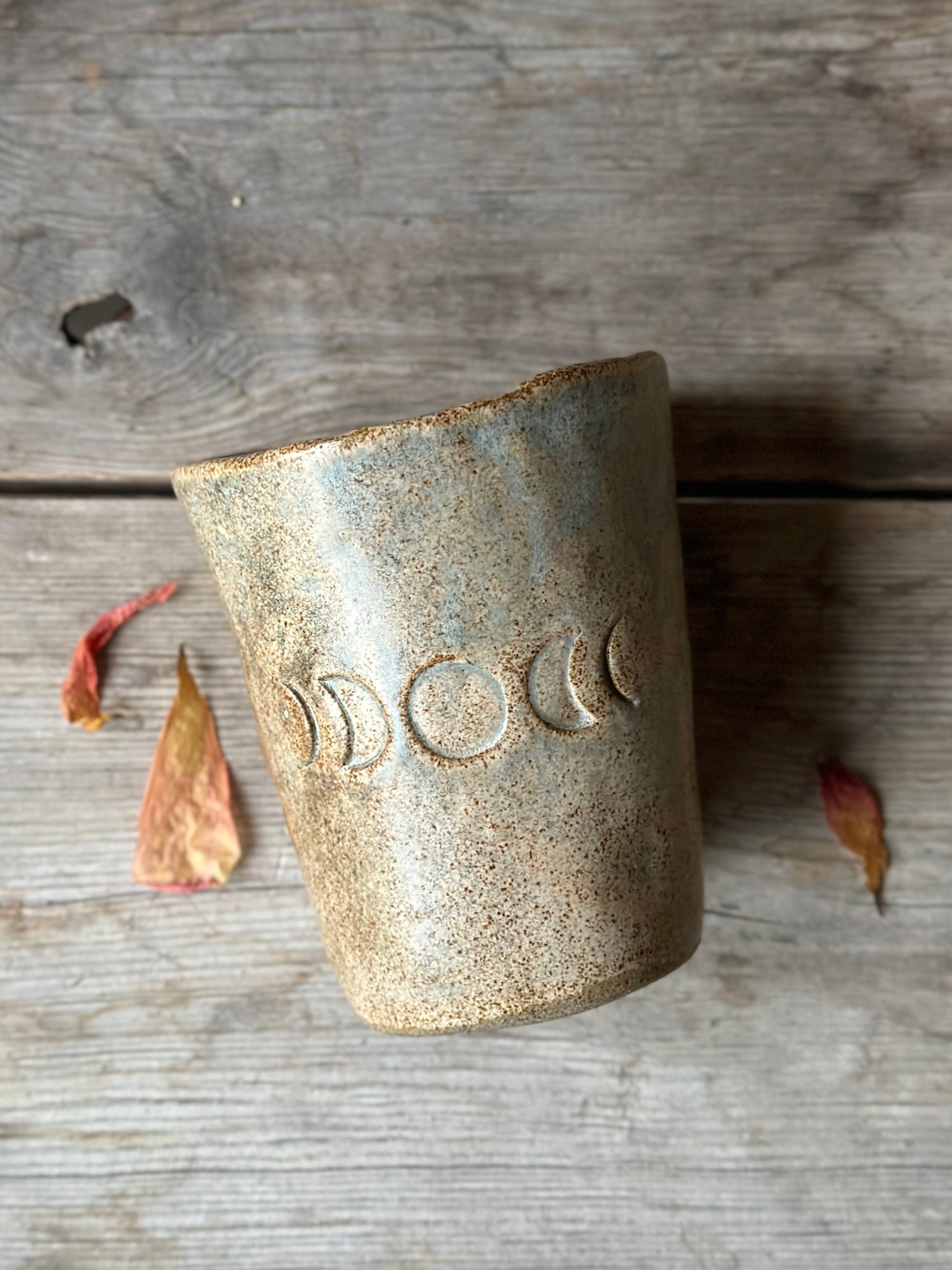 Beige grey ceramic cup laying on a grey old table. The cup is decorated with moon phases. Around the cup is dried flowers.